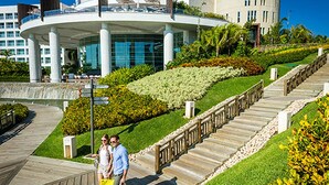 Un hotel moderno con fachada de vidrio curvo, un jardín ajardinado con diversas plantas y un paseo marítimo de madera que conduce a la entrada.