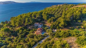 An aerial view of a coastal property with a large house, a swimming pool, and dense surrounding trees.