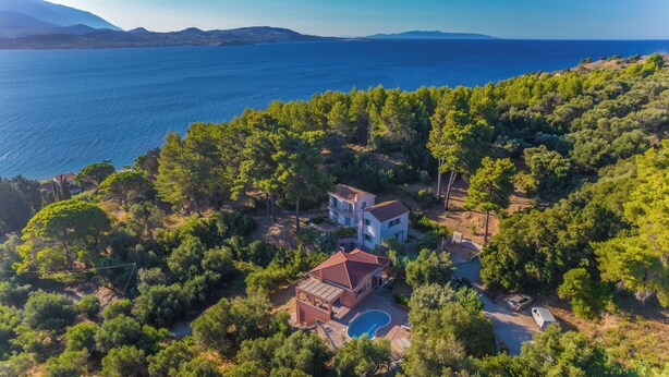 A two-story house with a swimming pool, surrounded by dense greenery and overlooking a large body of water.
