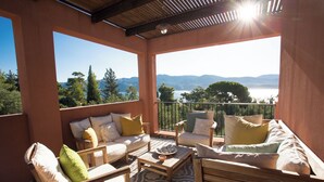 A sunlit balcony with a view of mountains and a lake, furnished with wooden chairs and a sofa.