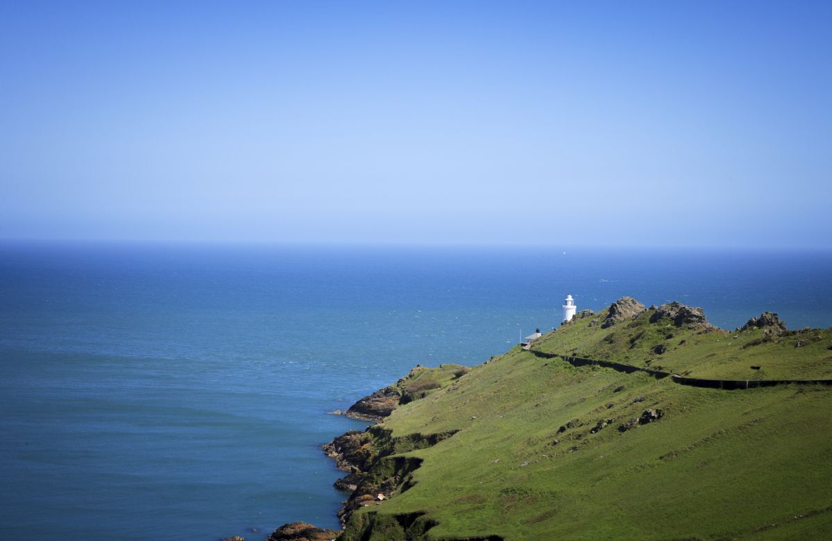 Eine Küstenlandschaft mit einem Leuchtturm auf einem grasbewachsen Hügel mit Blick aufs Meer.
