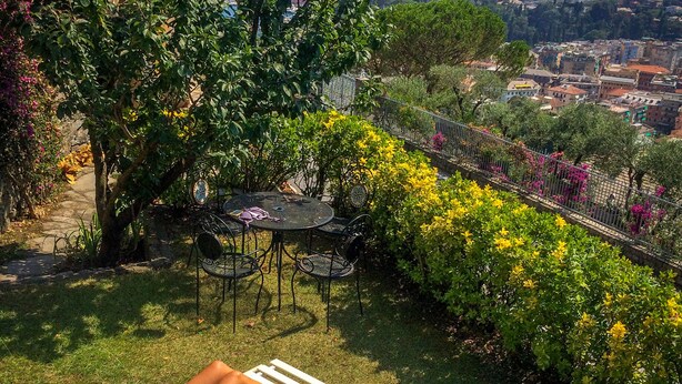 Une femme se détend sur un transat au bord d’une piscine, entourée d’une végétation luxuriante et avec une vue sur une ville perchée sur une colline.