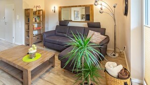 Una sala de estar con un sofá oscuro, una mesa de centro de madera, una planta en maceta, una estantería y una lámpara de pie.