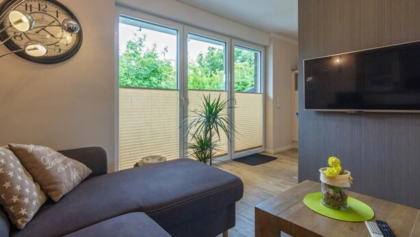 Una sala de estar moderna con un sofá, un reloj de pared, un televisor de pantalla plana y ventanas grandes con persianas.