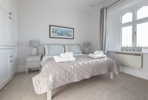 A bedroom with a bed, two bedside tables, a window with curtains, a radiator, and a framed picture on the wall.