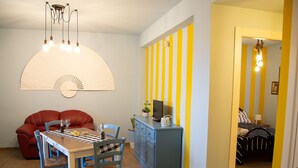 Et spiseområde med et bord, stole og en rød sofa. Der er et blåt skab og et fjernsyn på et stativ.