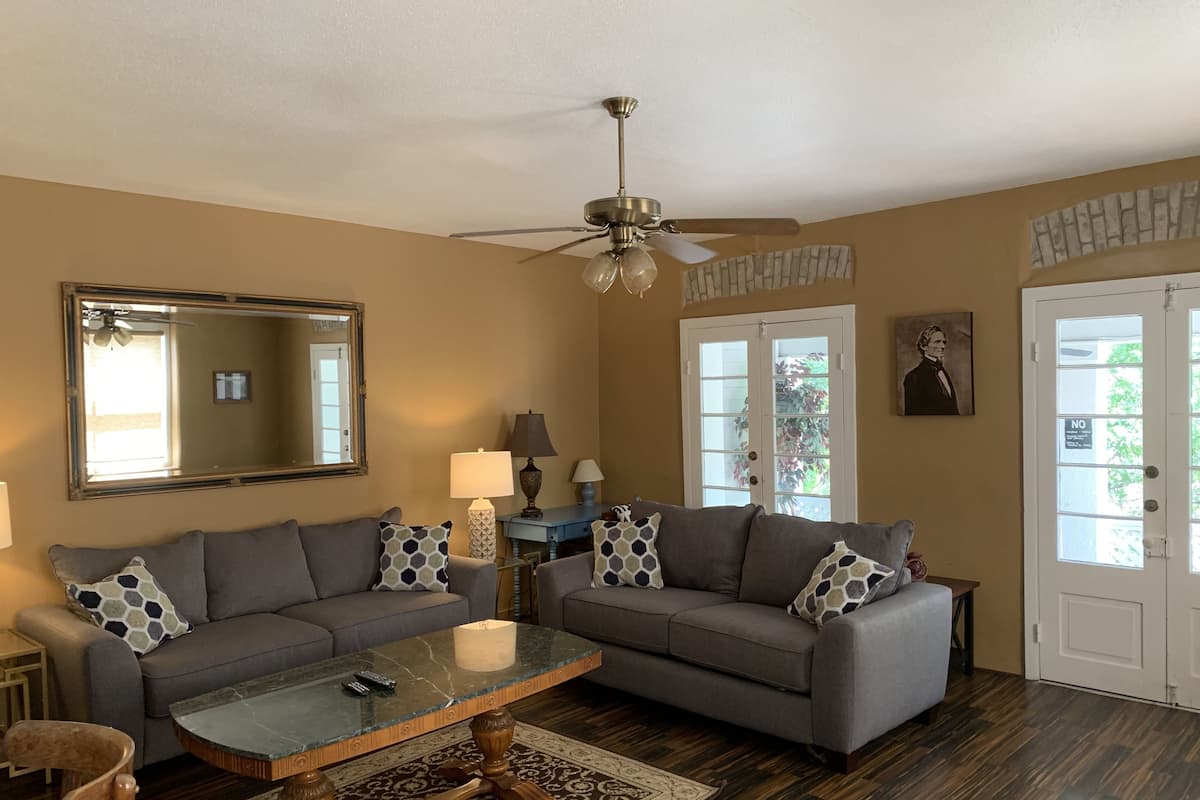 A living room with two grey sofas, a glass coffee table, a wooden chair, a ceiling fan, and a large mirror.