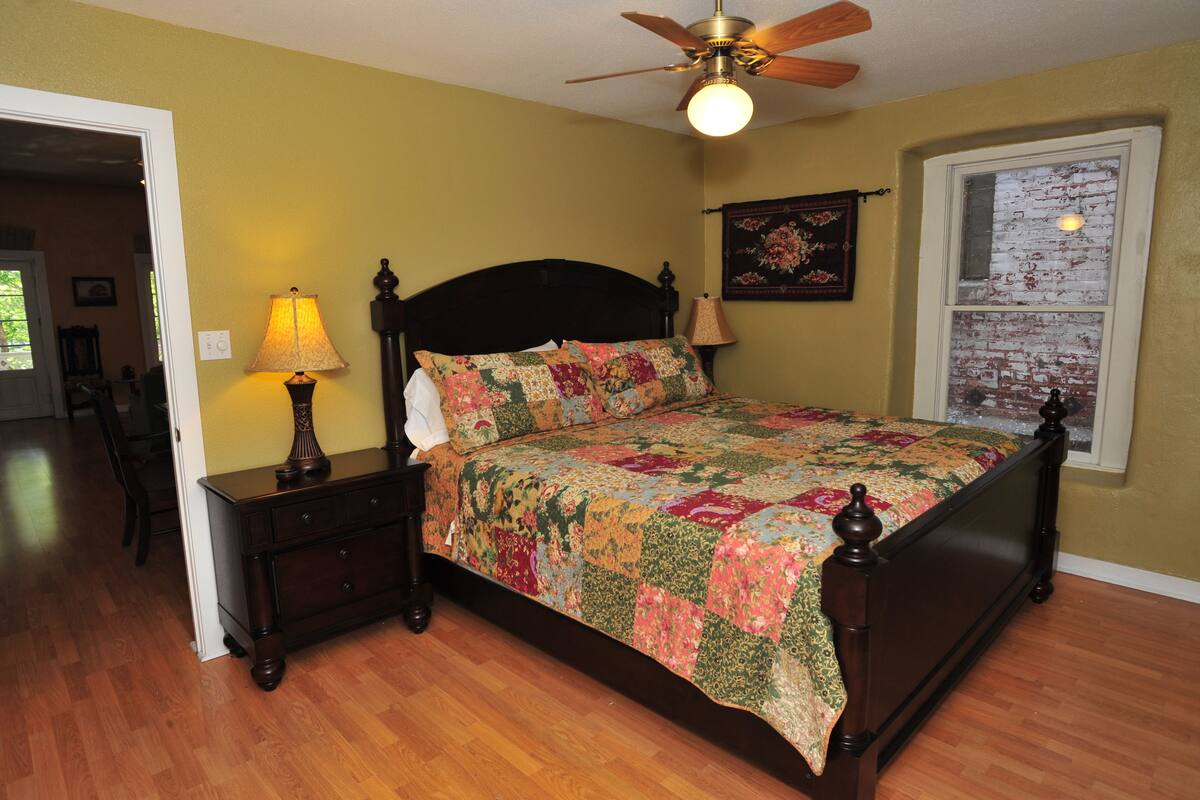 A bedroom with a wooden bed, a nightstand with a lamp, a ceiling fan, and a window with a view of the outside.