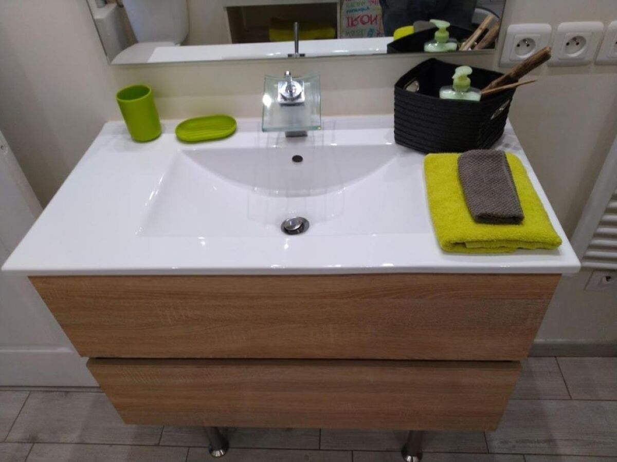 Un baño con un lavamanos blanco, cajones de madera, una taza verde, una toalla amarilla y un jabonera negra.