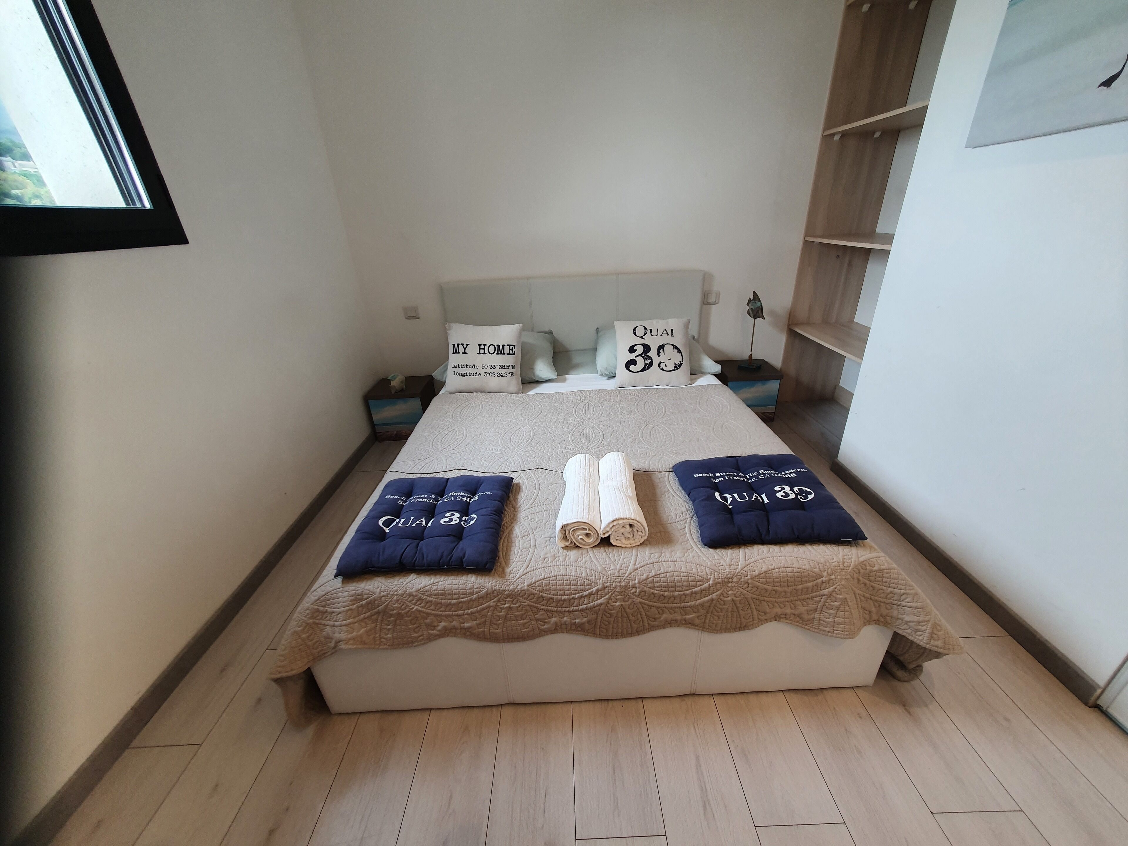 A neatly made bed with a quilted headboard, two rolled towels, and two pillows with 'Quai 30' text.