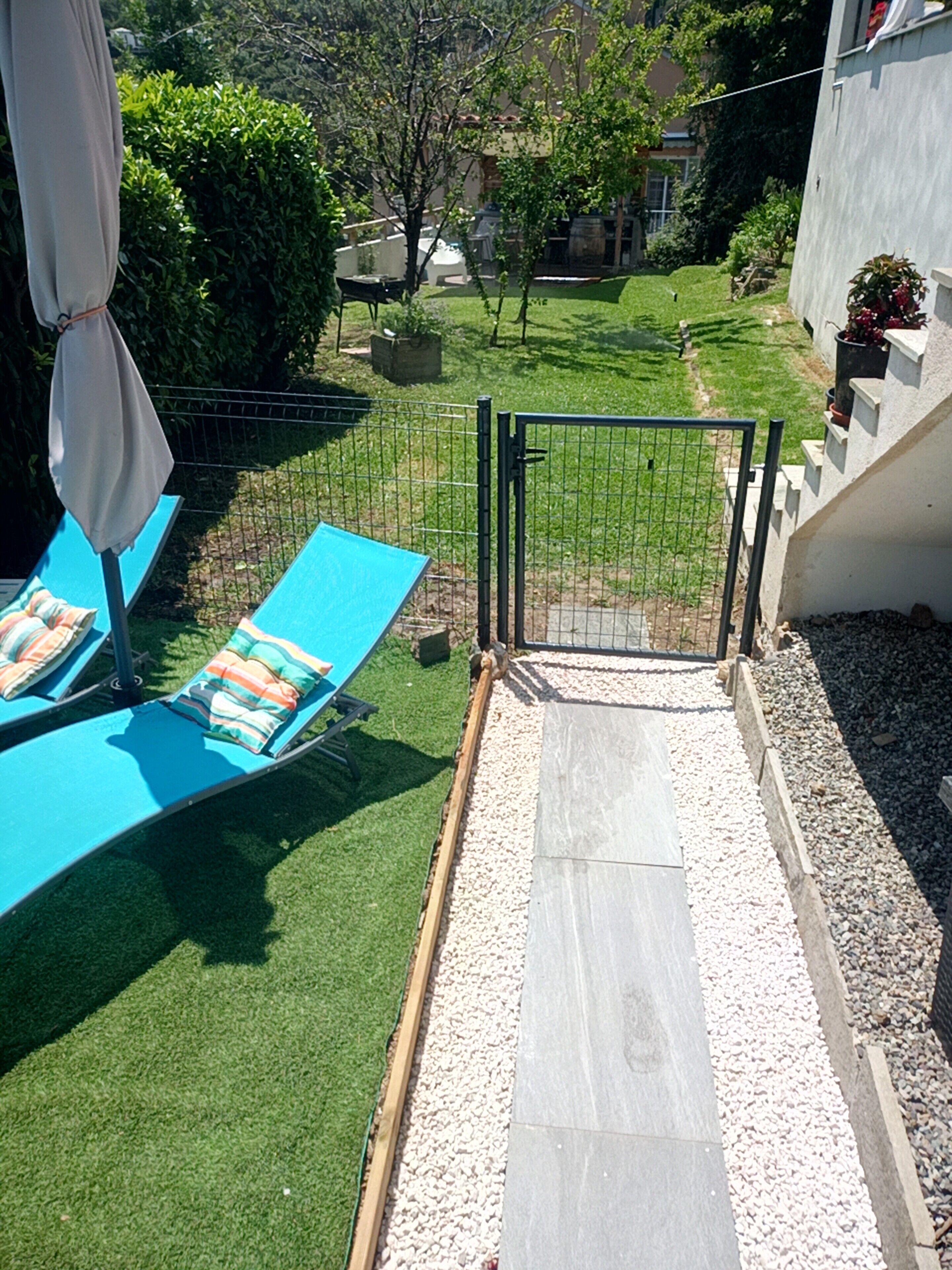 Un jardin avec un fauteuil de relaxation bleu, un petit potager, un portail en métal et un chemin pavé.