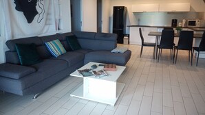 A modern living room with a grey sofa, a coffee table with magazines, and a kitchen area with white cabinets and a black refrigerator. - Uribellinu 2, nice T3 mountain sea view (santa maria di lota)