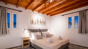 Een slaapkamer met een bed, houten balken in het plafond, een nachtkastje, een aan de muur hangend schilderij en een raam met gordijnen.