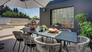 En udendørs terrasse med et langt bord, fletstole og en stor parasol.