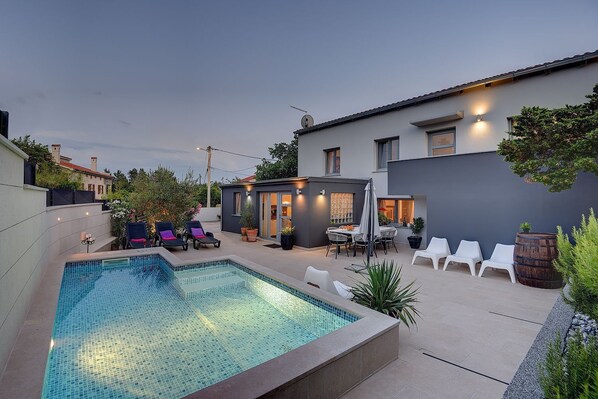 A modern outdoor pool area with lounge chairs, a dining table, and a small garden.