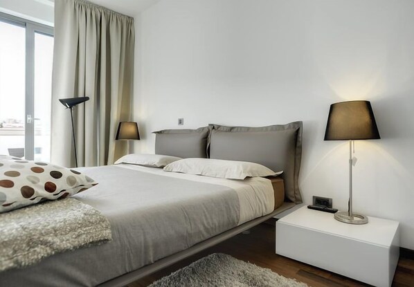 Ein modernes Schlafzimmer mit einem Bett, zwei Nachttischlampen, einem Nachttisch und Blick auf die Stadt durch große Fenster.