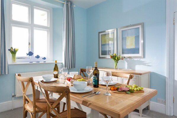 A dining area with a wooden table set for four, surrounded by chairs. There are plates, bowls, cutlery, and a bottle of wine on the table.