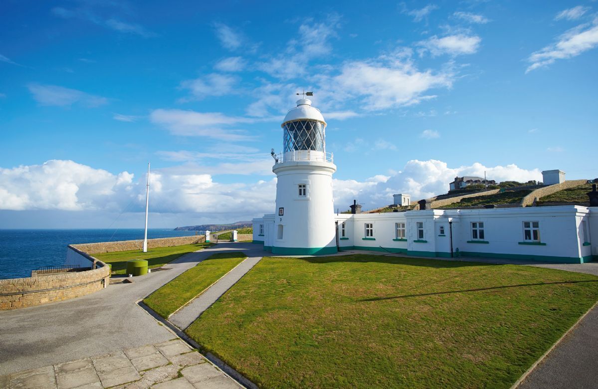A white lighthouse with a glass lantern room, surrounded by a grassy area and a stone wall, with the sea in the background.