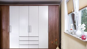 Ein Zimmer mit großem Kleiderschrank, einem Fenster mit Jalousien und einer Vase mit Blumen auf der Fensterbank.