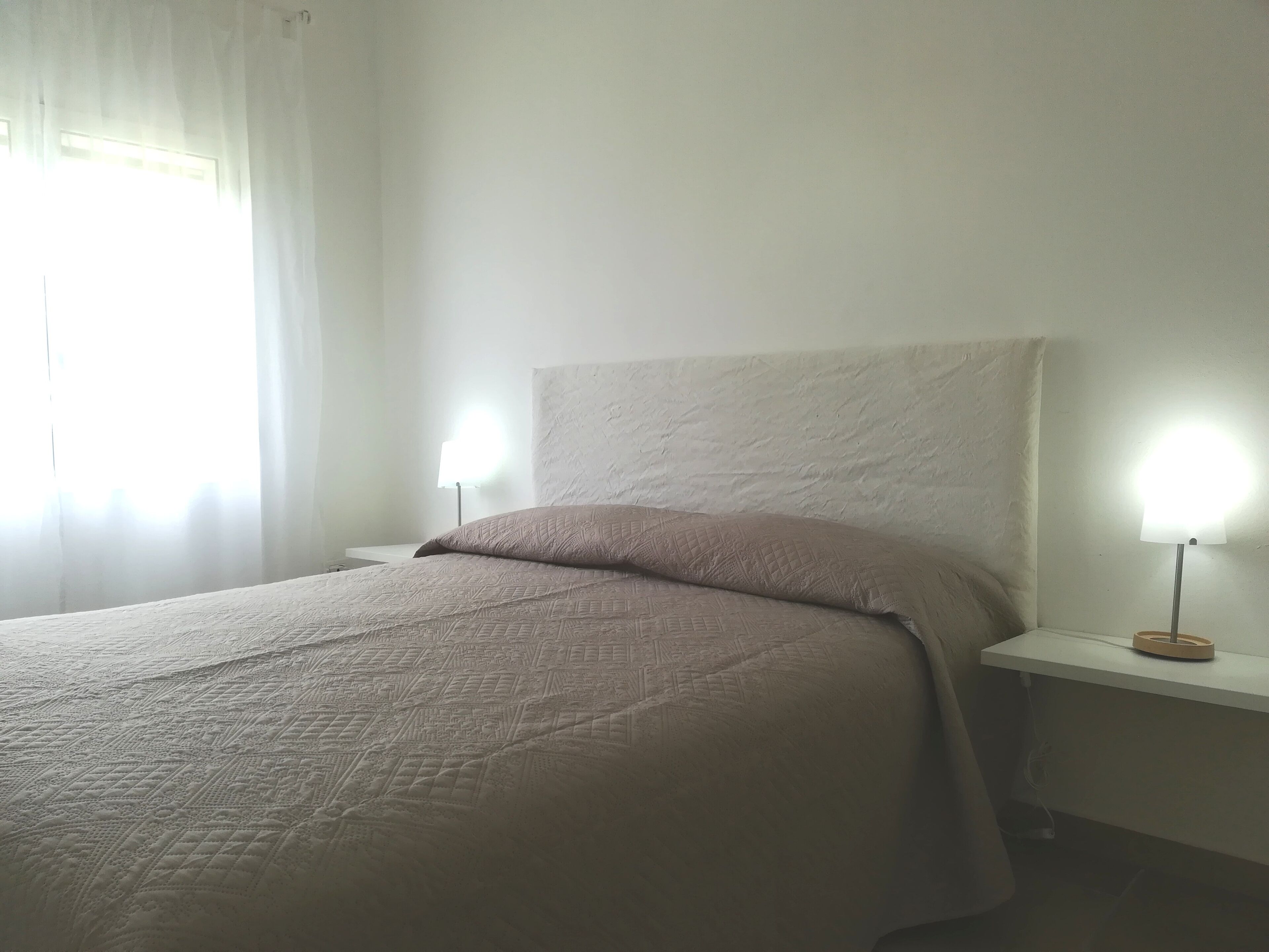 Een keurig opgemaakt bed met een bedsprei met patroon, twee bedlampen en een raam met dunne gordijnen.