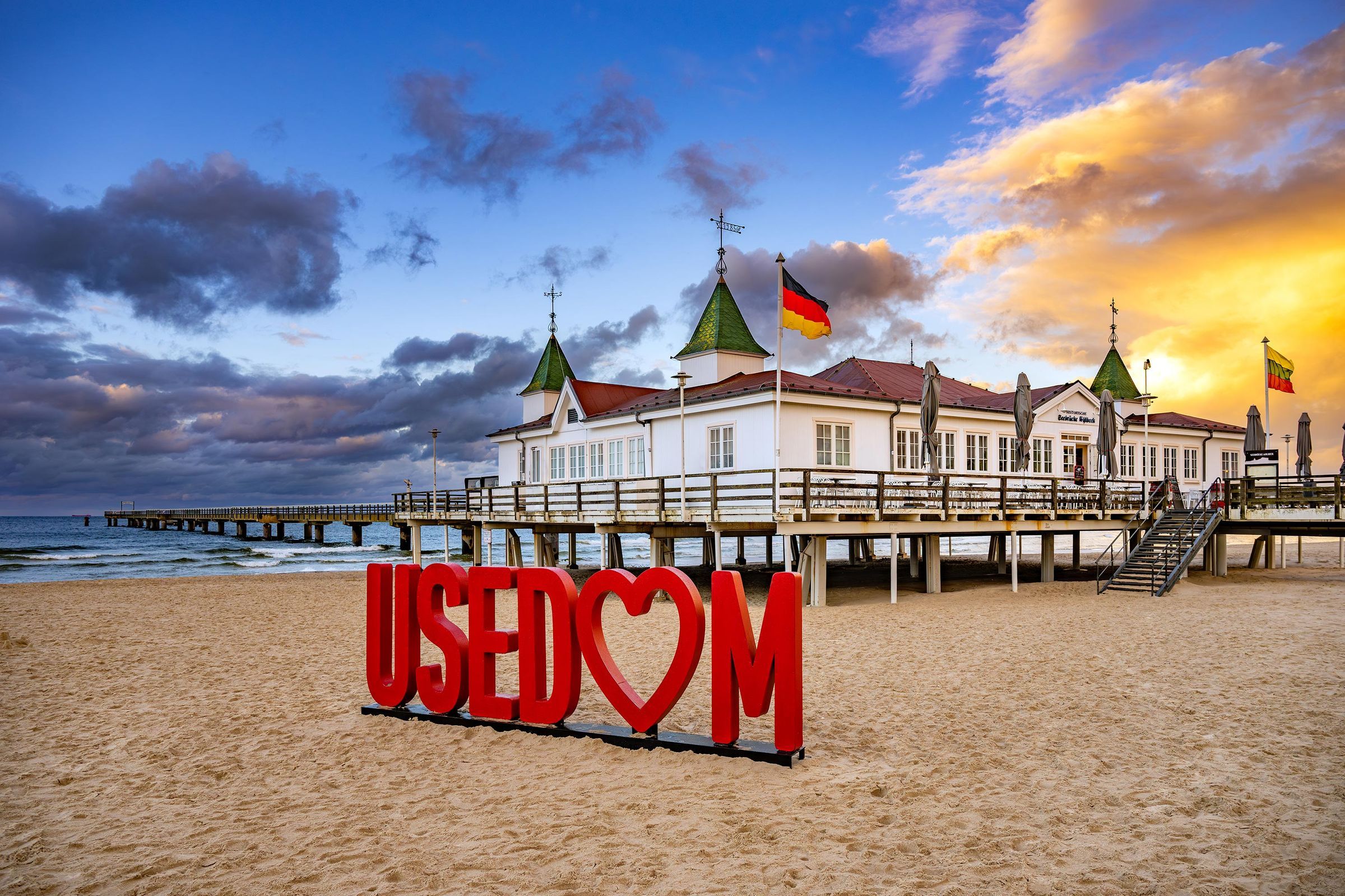 Una playa con un cartel grande y rojo que dice 'USEDOM' y un edificio con varias banderas.