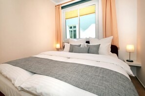 Ein ordentlich bezogenes Bett mit einer grau-weißen Decke, zwei Nachttischlampen und einem Fenster mit Vorhängen.