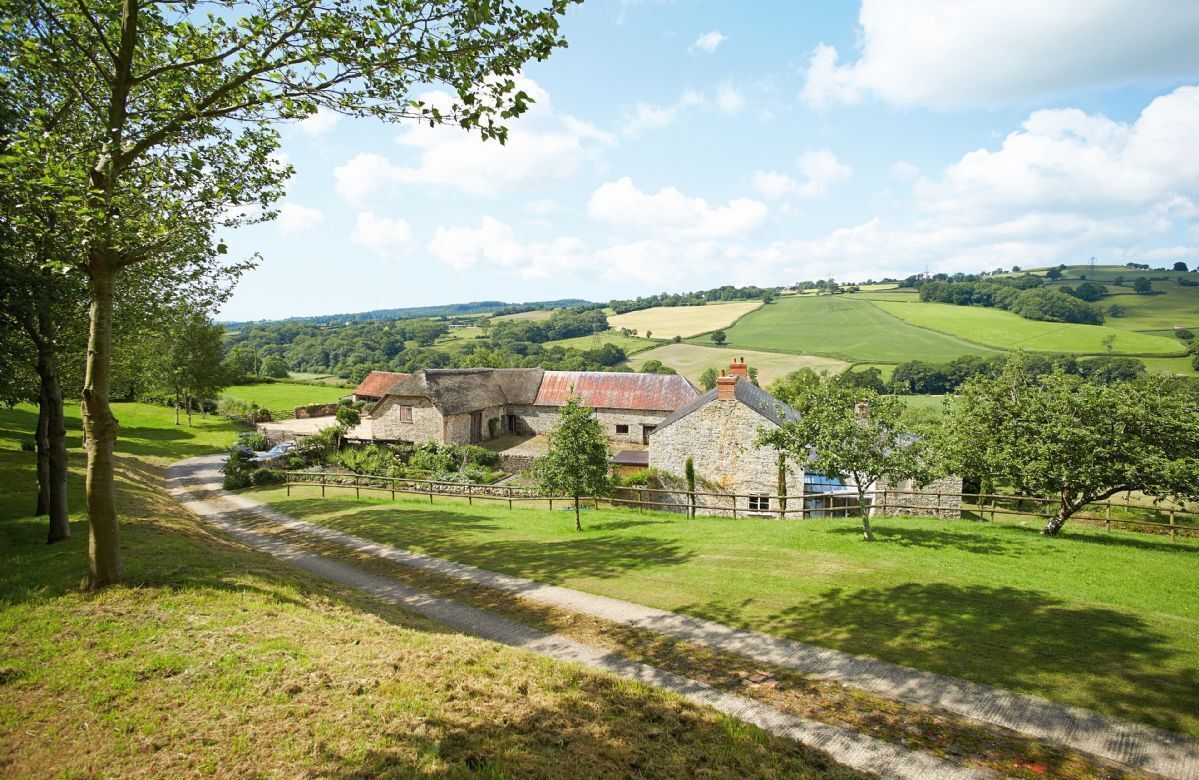 A stone farmhouse with a thatched roof, surrounded by lush green fields and a clear sky.