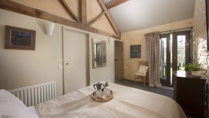 Ein Schlafzimmer mit einem großen Bett, einer Holzbalkendecke, einem Heizkörper, einem Nachttisch mit einer Tablett voller Tassen und einer Blume, und einer Tür zu einem Balkon.