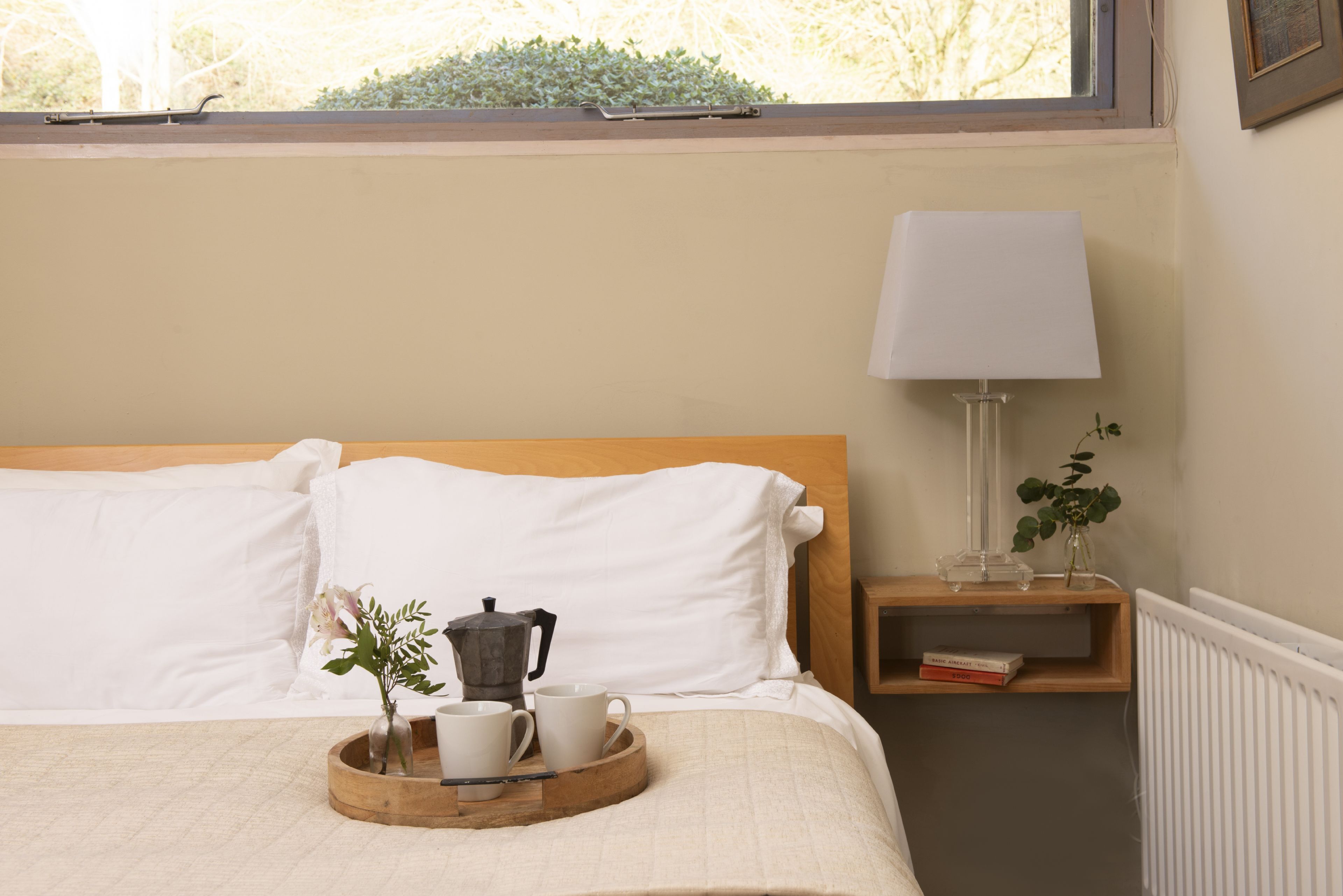 A neatly made bed with white linens, a bedside table with a lamp, a tray with a coffee pot and cups, and a small plant.