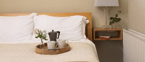 A neatly made bed with white linens, a bedside table with a lamp, a tray with a coffee pot and cups, and a small plant.