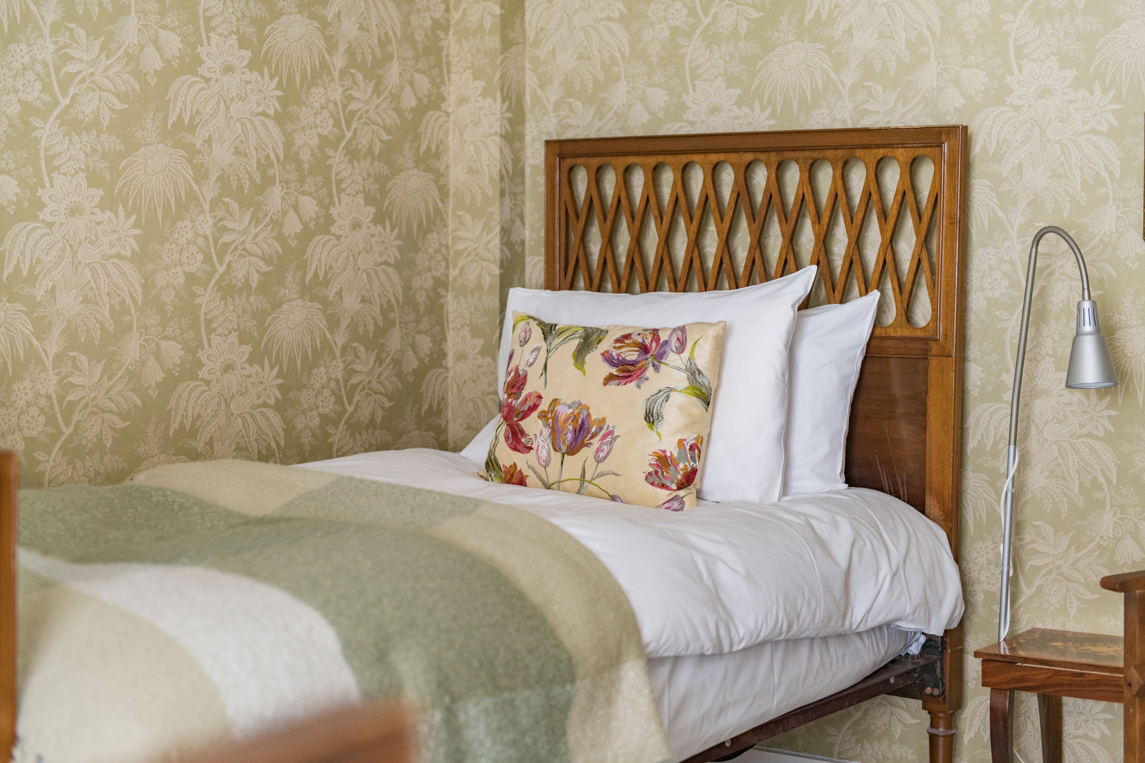A neatly made bed with a floral pillow and a wooden headboard, a bedside table with a lamp, and floral wallpaper.