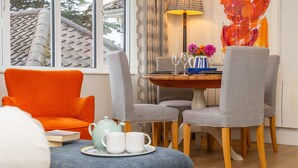 A room with a dining table, chairs, a sofa, a lamp, and a window with a view of a tiled roof. - River View, Aldeburgh (Aldeburgh)