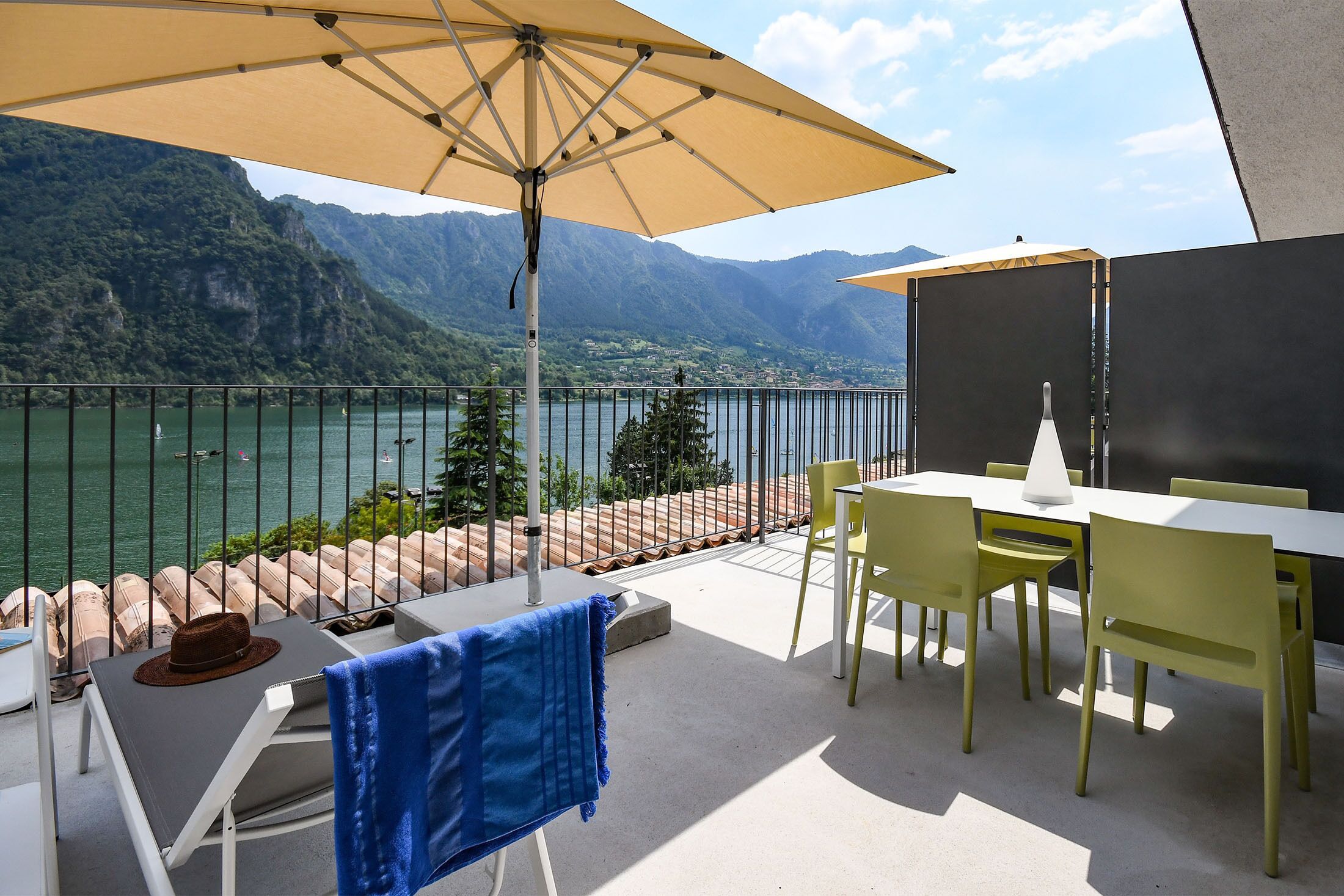 Ein Balkon mit Tisch und Stühlen, einem gelben Sonnenschirm und Blick auf einen See und Berge.