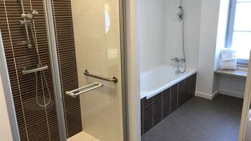 A modern bathroom with a glass-enclosed shower, a bathtub, and a window with towels.