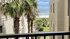 A balcony with a view of the ocean, palm trees, and a building with a flat roof.