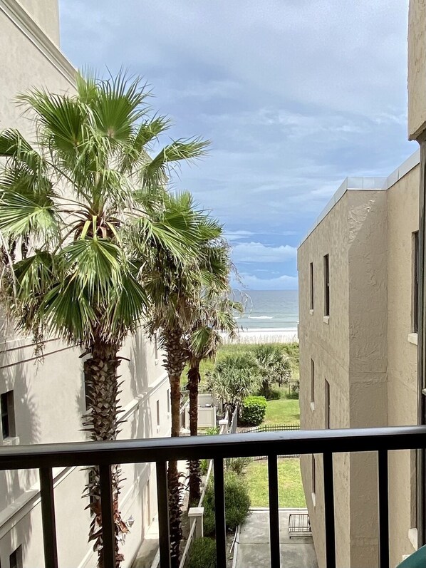 A balcony with a view of the ocean, palm trees, and a building with a flat roof.