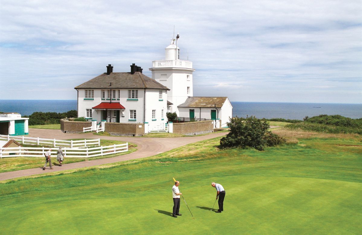 A coastal building with a lighthouse, a golf course, and a white fence.