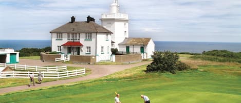 A coastal building with a lighthouse, a golf course, and a white fence.