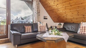 Ein modernes Wohnzimmer mit Holzboden, einer Sofagarnitur, einem Couchtisch mit Blumenarrangement und Blick auf eine gebirgige Landschaft durch ein großes Fenster.