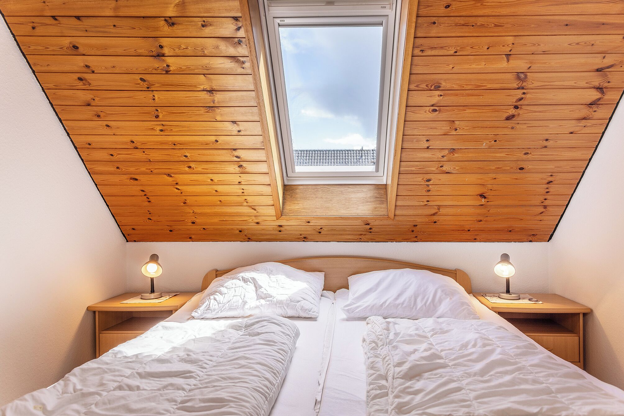 Une chambre avec un plafond en pente, un puits de lumière, deux lampes de chevet et un lit avec une literie blanche.