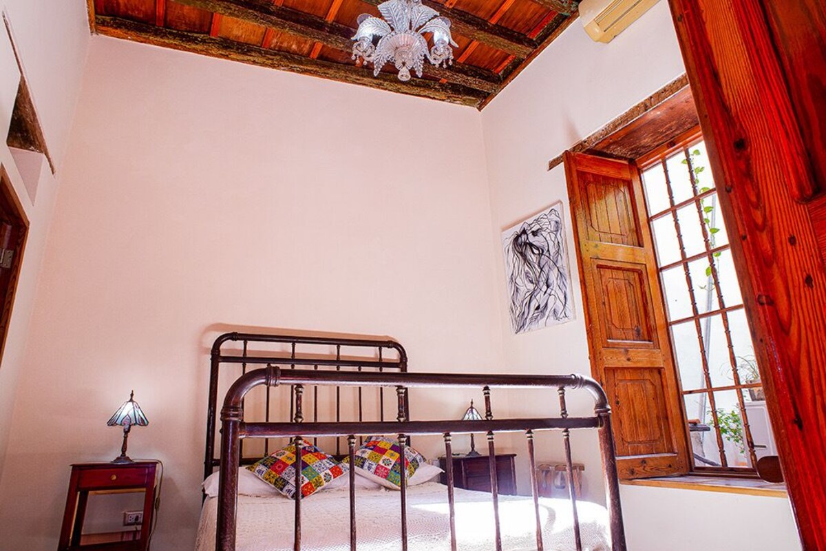 A bedroom with a wooden bed, a nightstand, a wall-mounted lamp, and a window with a view.
