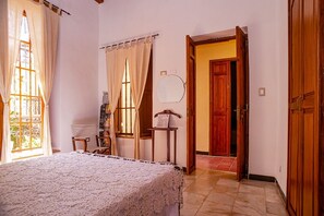 Un dormitorio con cama, ventana con cortinas, espejo y puerta de madera que da a otra habitación.