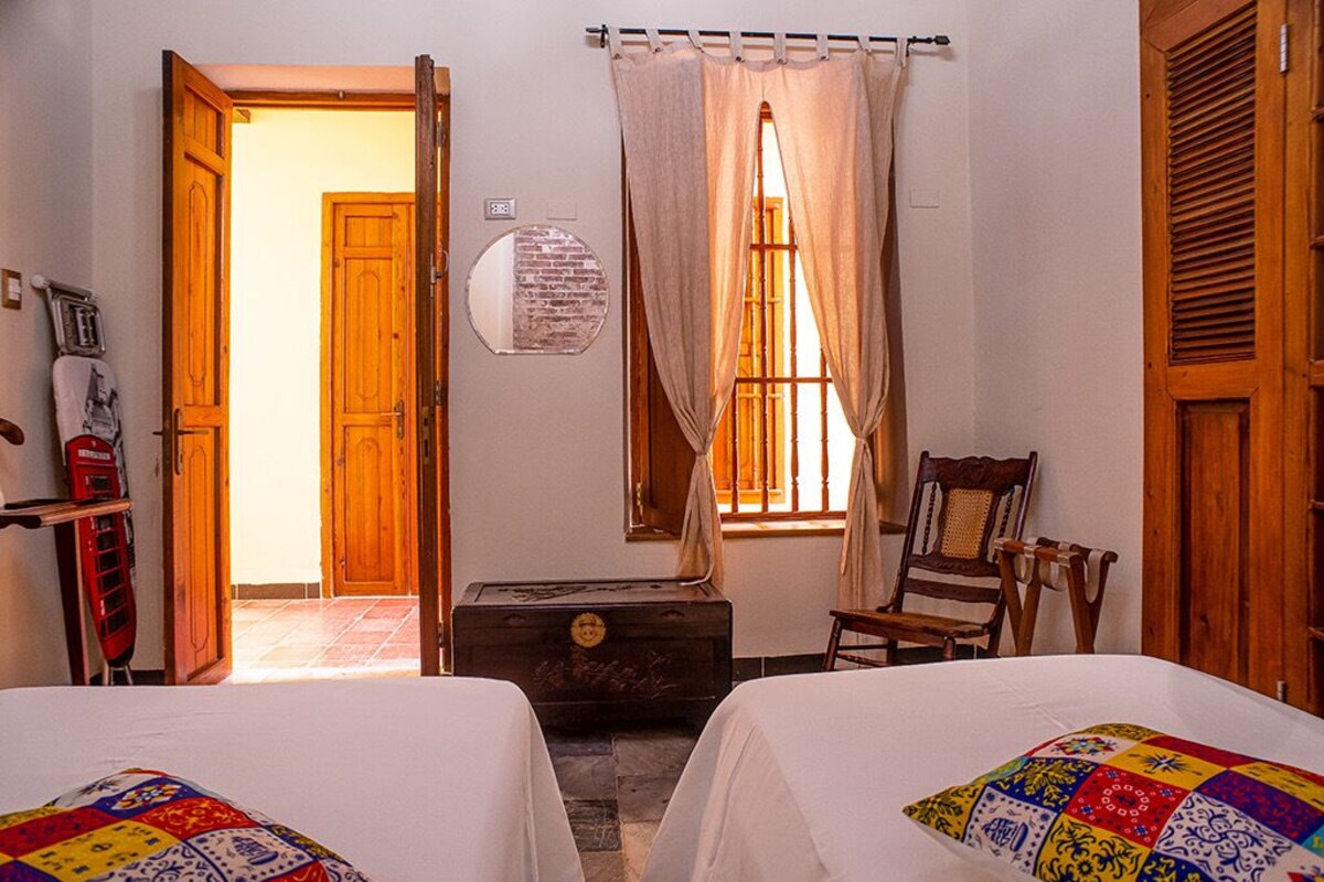 A room with two beds, a wooden chest, a chair, a wooden door, and a window with curtains.