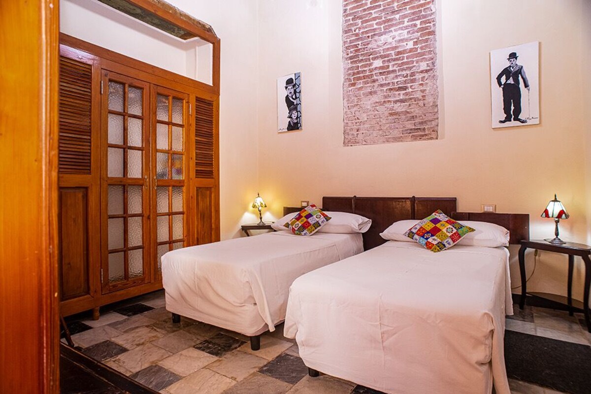 A hotel room with two single beds, a wooden wardrobe with glass doors, a brick wall, and two wall-mounted lamps.