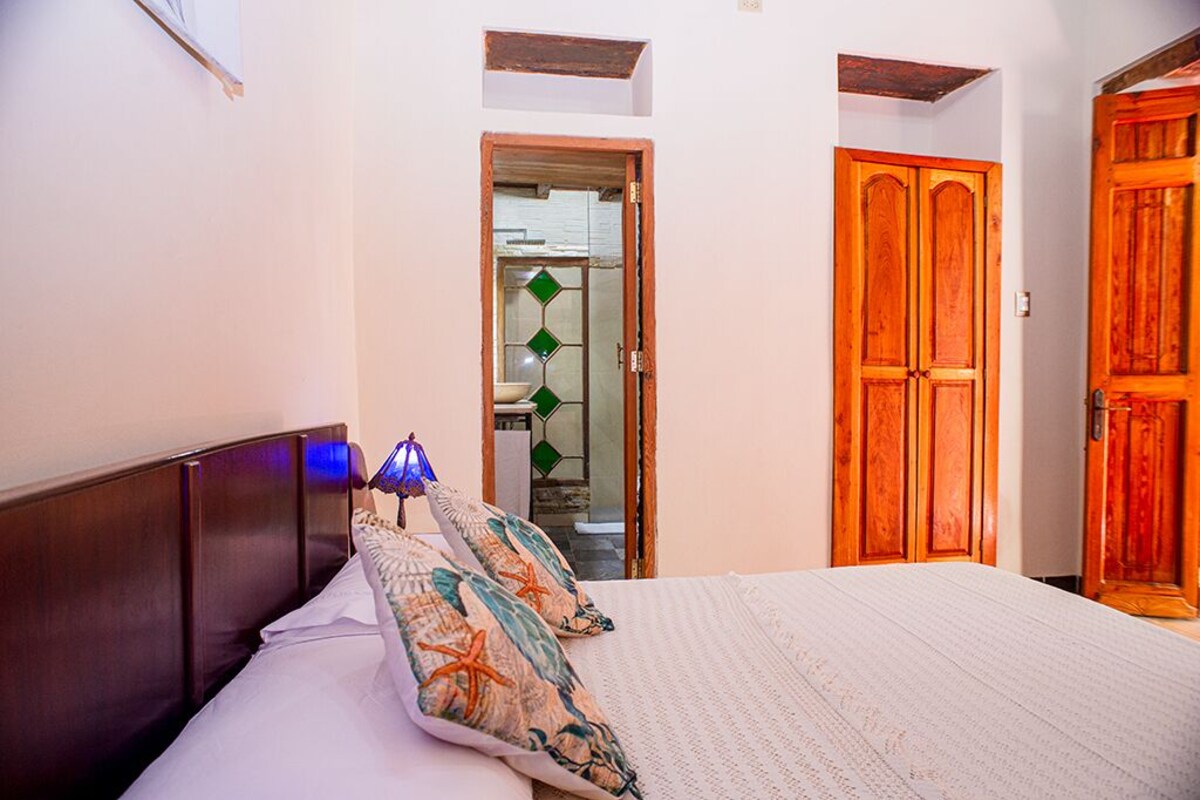 A bedroom with a wooden headboard, a bed with patterned pillows, a nightstand with a lamp, and a wooden wardrobe.