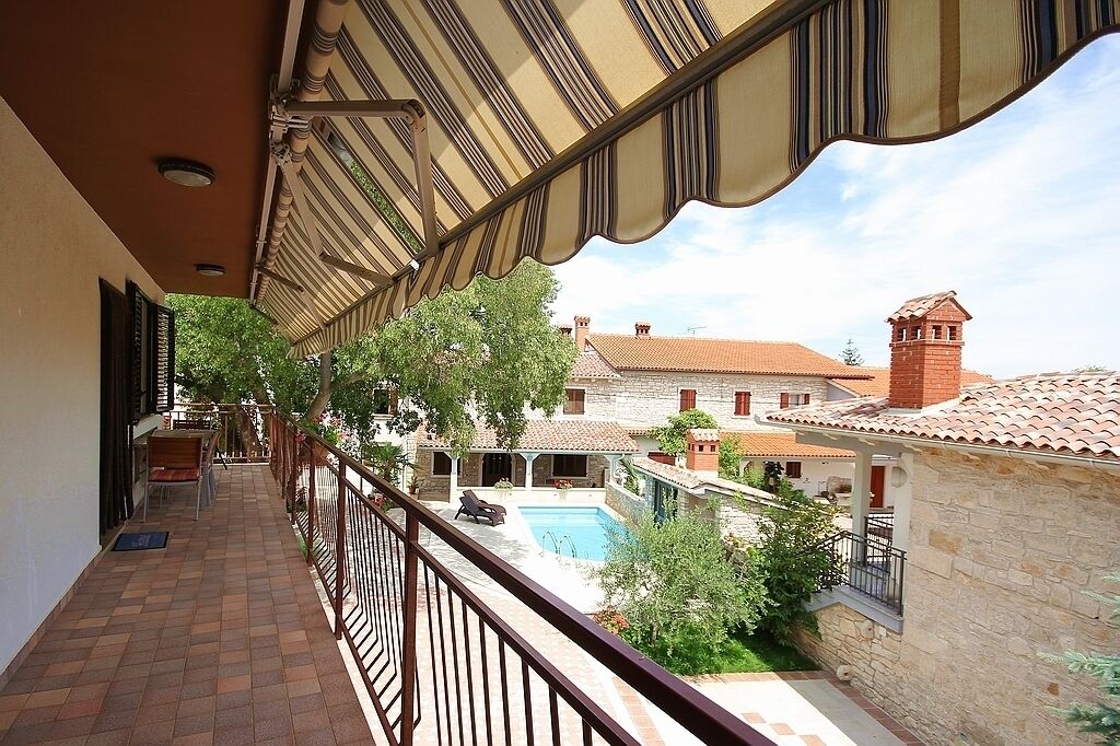 Un balcon donnant sur une piscine et une cour, avec en toile de fond un bâtiment en pierre doté d’un toit recouvert de tuiles rouges et d’une tour.