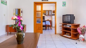 Uma sala de estar com uma mesa de jantar de madeira, uma TV sobre um suporte de madeira e uma cozinha ao fundo.