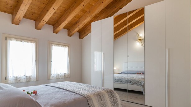 Een slaapkamer met een houten plafond, een bed met een wit en patroonrijk beddegoed, en een grote spiegel die een ander bed reflecteert.