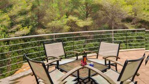Una terraza con cuatro sillas y una mesa, rodeada por una barandilla metálica, con vistas a un bosque verde y frondoso.