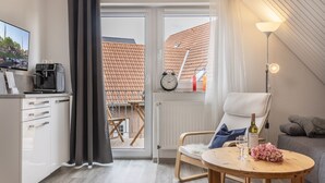 A modern room with a balcony, a coffee machine, a chair, a table with a bottle and flowers, and a TV on the left.